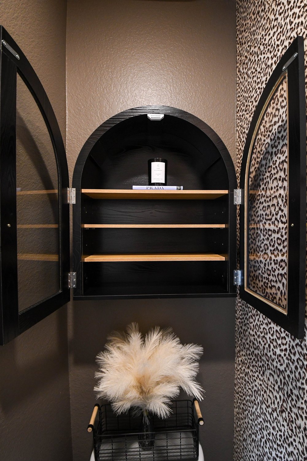 Arched black cabinet opened to show its wood shelves against the leopard wallpaper