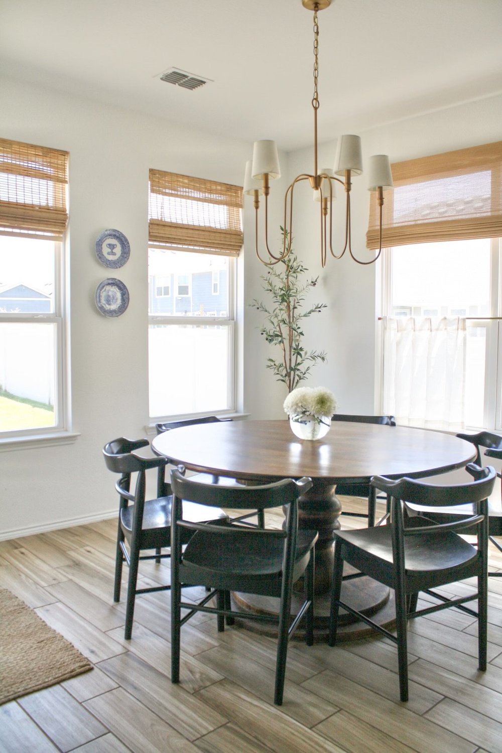 Round wood dining table and black spindle chairs under a brass chandelier in a bay window