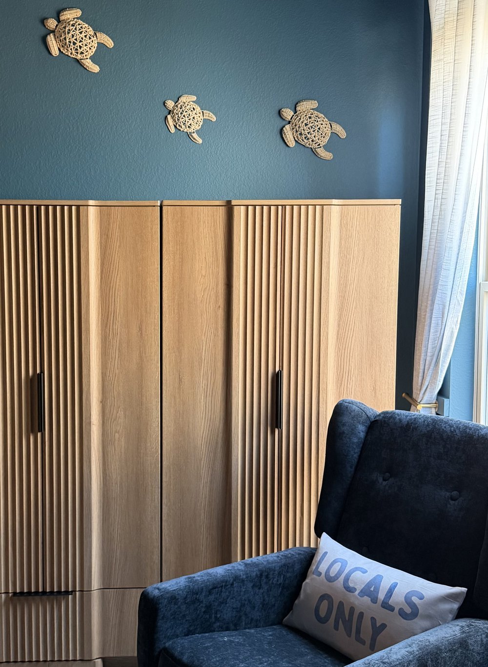 Fluted oak wardrobe beside a navy glider chair, carved turtles on the teal wall