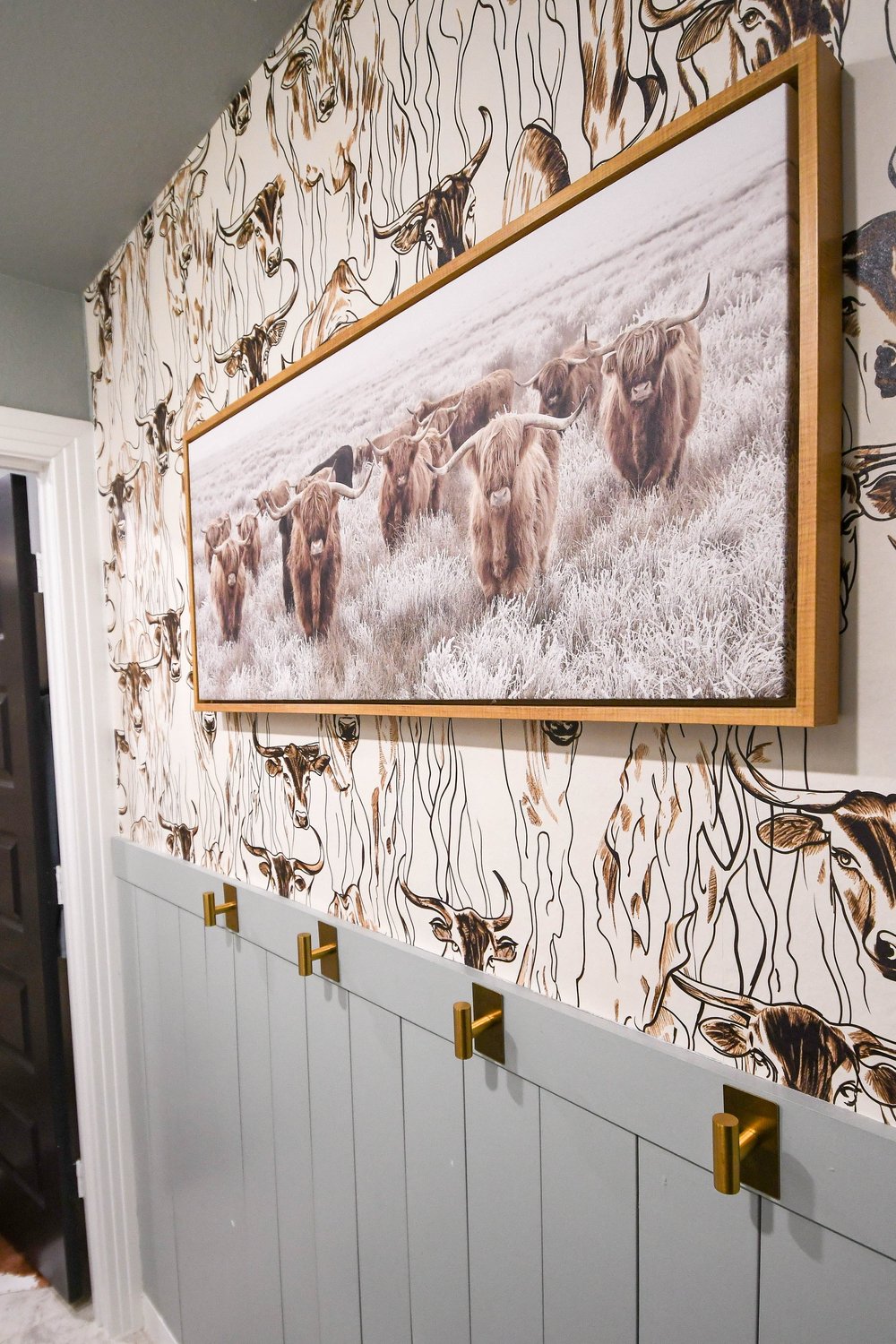Framed cattle canvas on the longhorn wallpaper above the sage wainscot with brass hooks