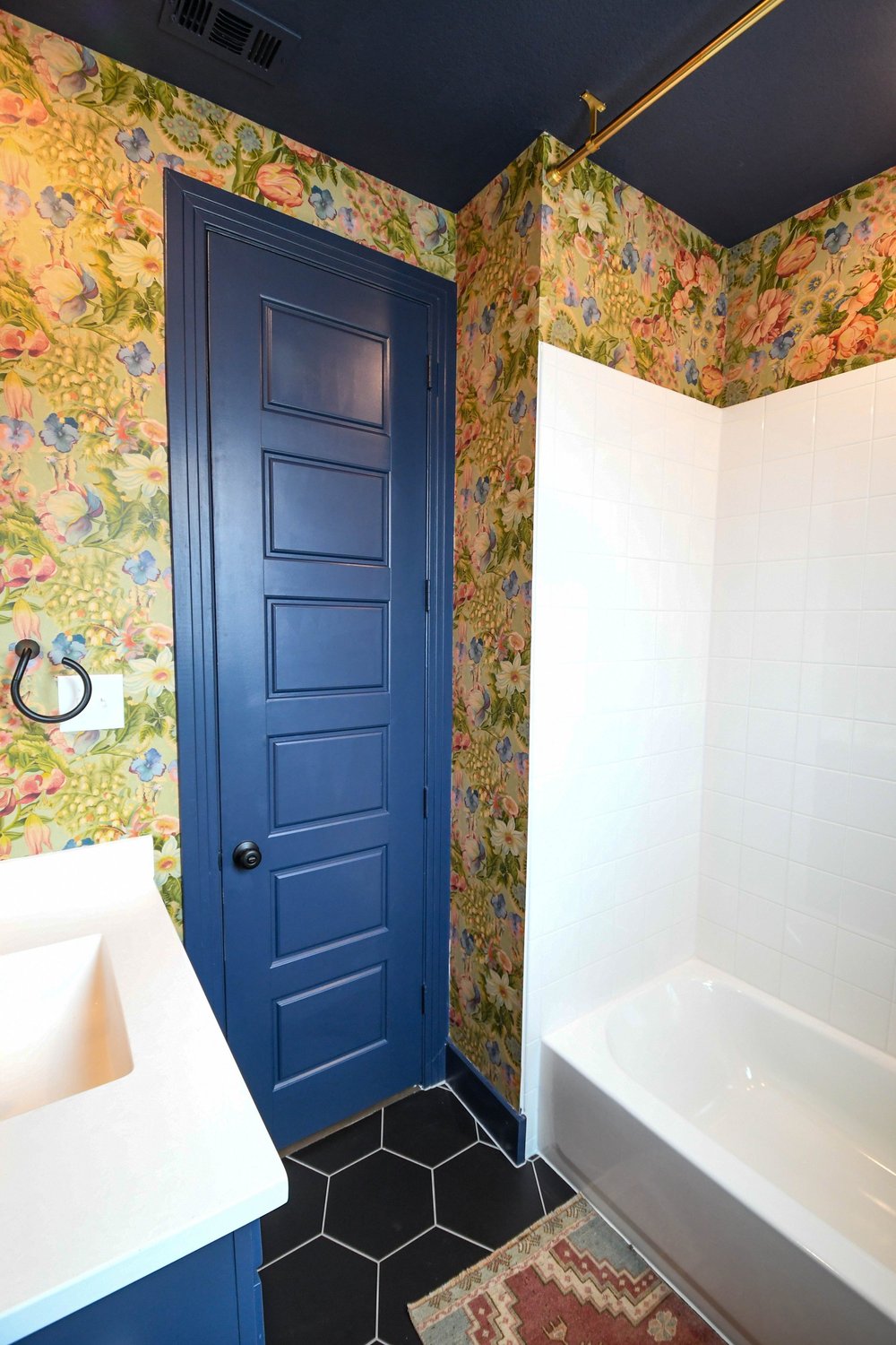Navy door and white tub shower next to the vanity in the wallpapered bathroom