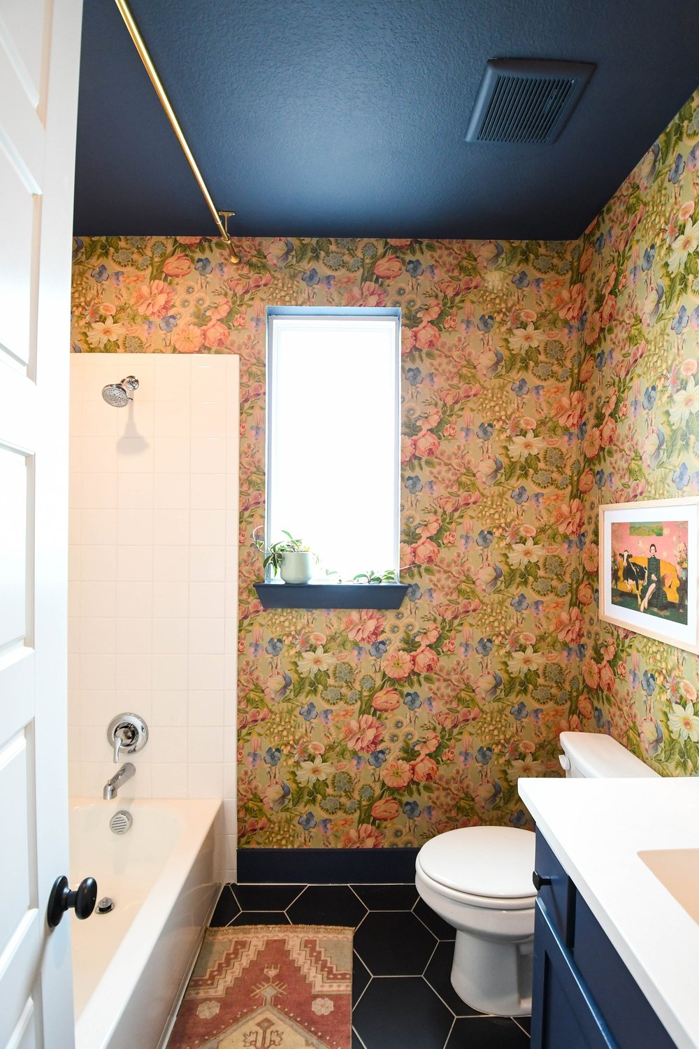 The tub shower, window and navy vanity with the wallpaper carried to the ceiling