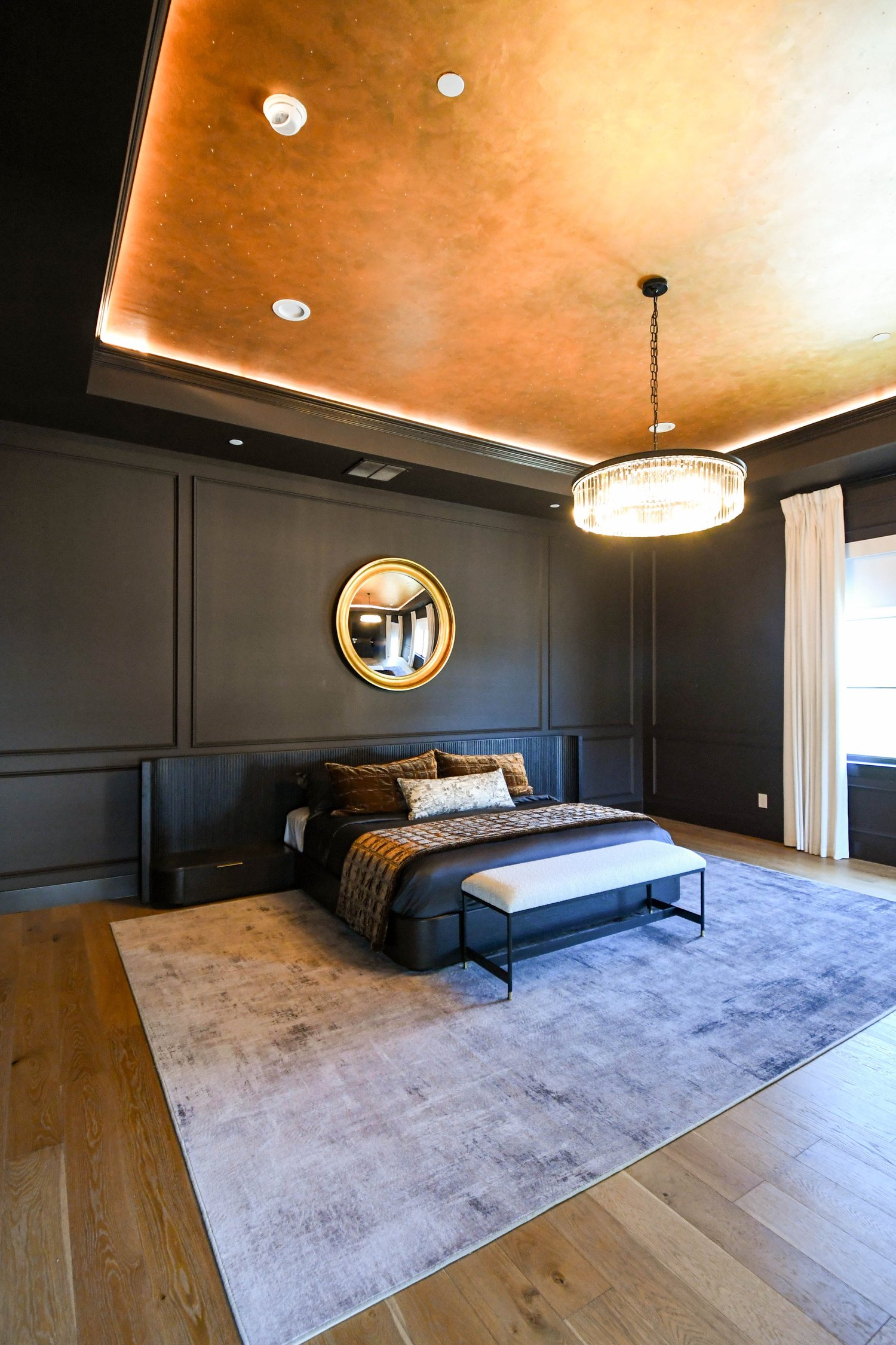 A dark panelled primary bedroom with a gold-framed mirror, a warm gold-lit tray ceiling and a crystal chandelier