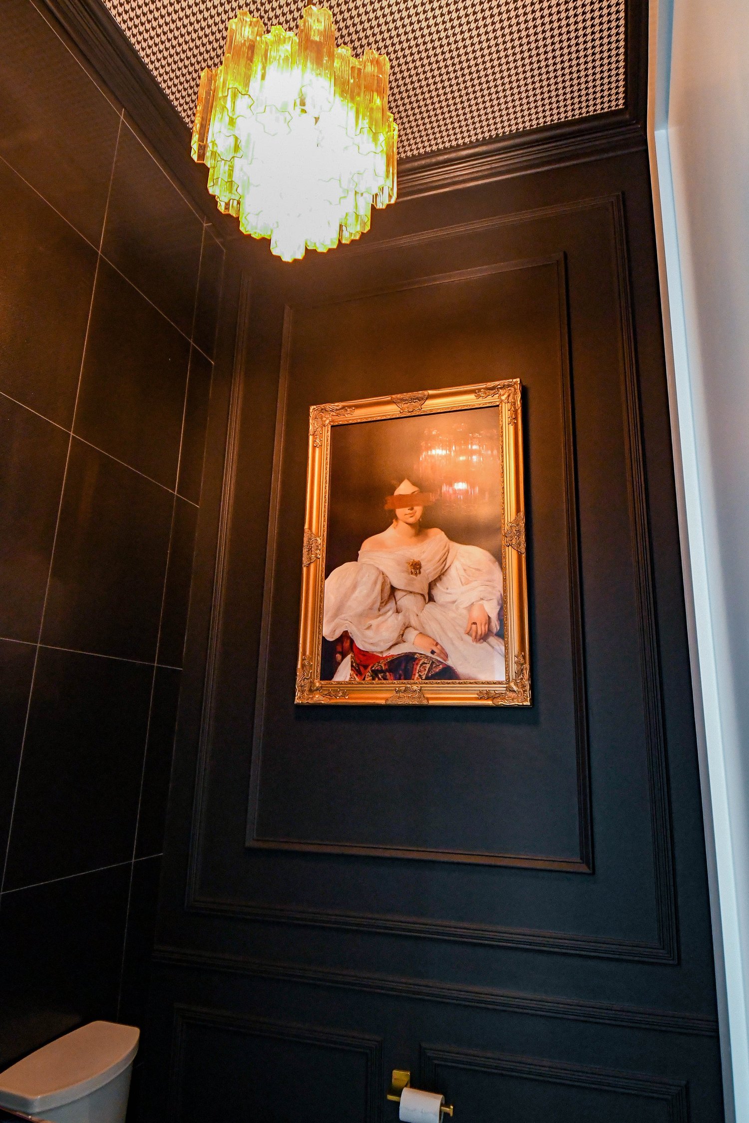 A powder bath with dark panelled walls, a framed portrait and a yellow glass chandelier